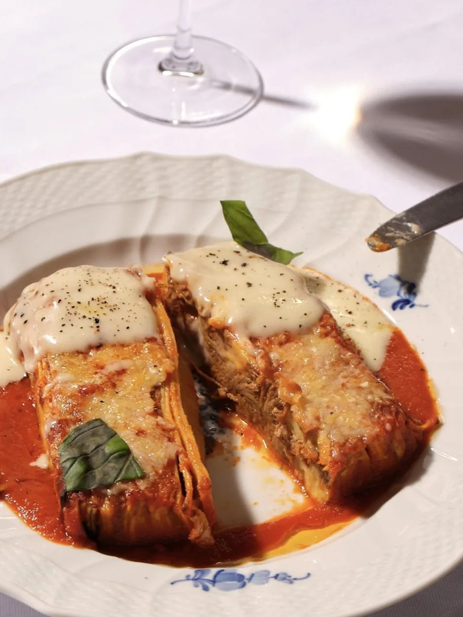 Baked pasta split in half on a blue floral porcelain plate, topped with béchamel, black pepper and basil, over tomato sauce.