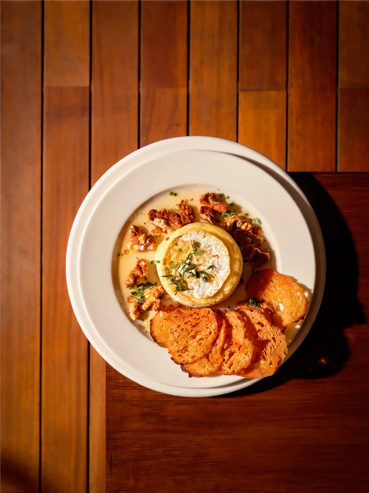 Baked camembert in a white dish on a wooden table, topped with thyme and surrounded by caramelized walnuts and toasted bread slices.