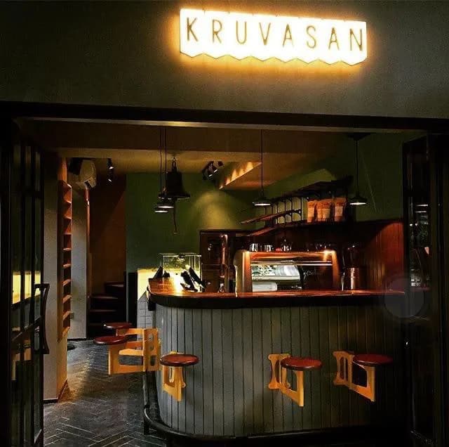 The first shop at night: an illuminated Kruvasan sign, green walls, a wooden counter, orange stools and a staircase to the upper floor.