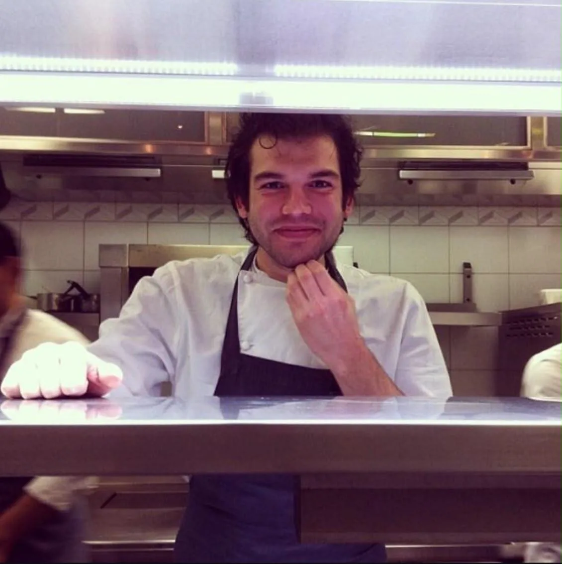 A young cook in a white chef's jacket and dark apron standing behind a steel service counter in a white-tiled kitchen.