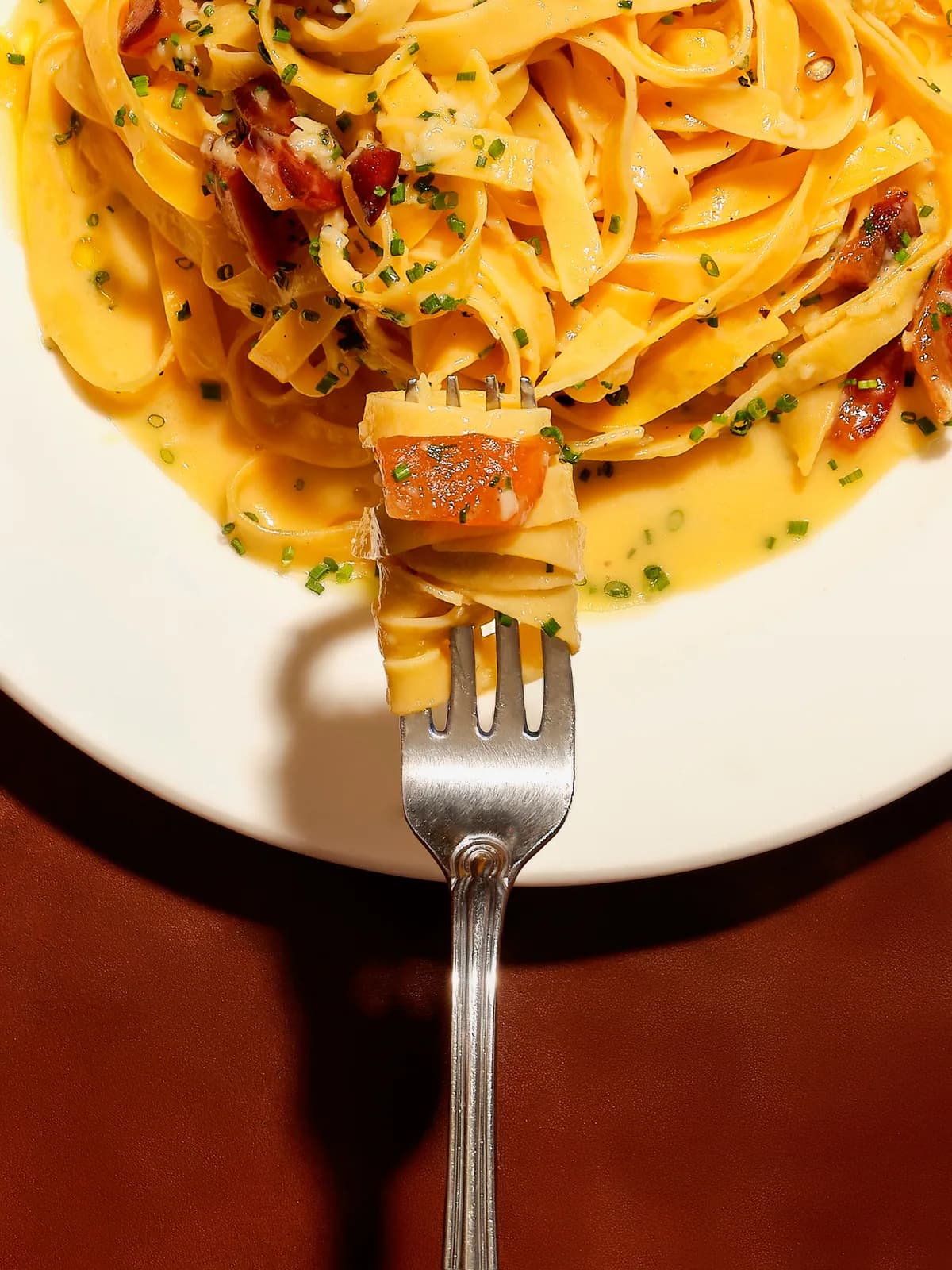 Creamy carbonara on a white plate; a fork lifting a bite, with crisped guanciale and chives mixed through.