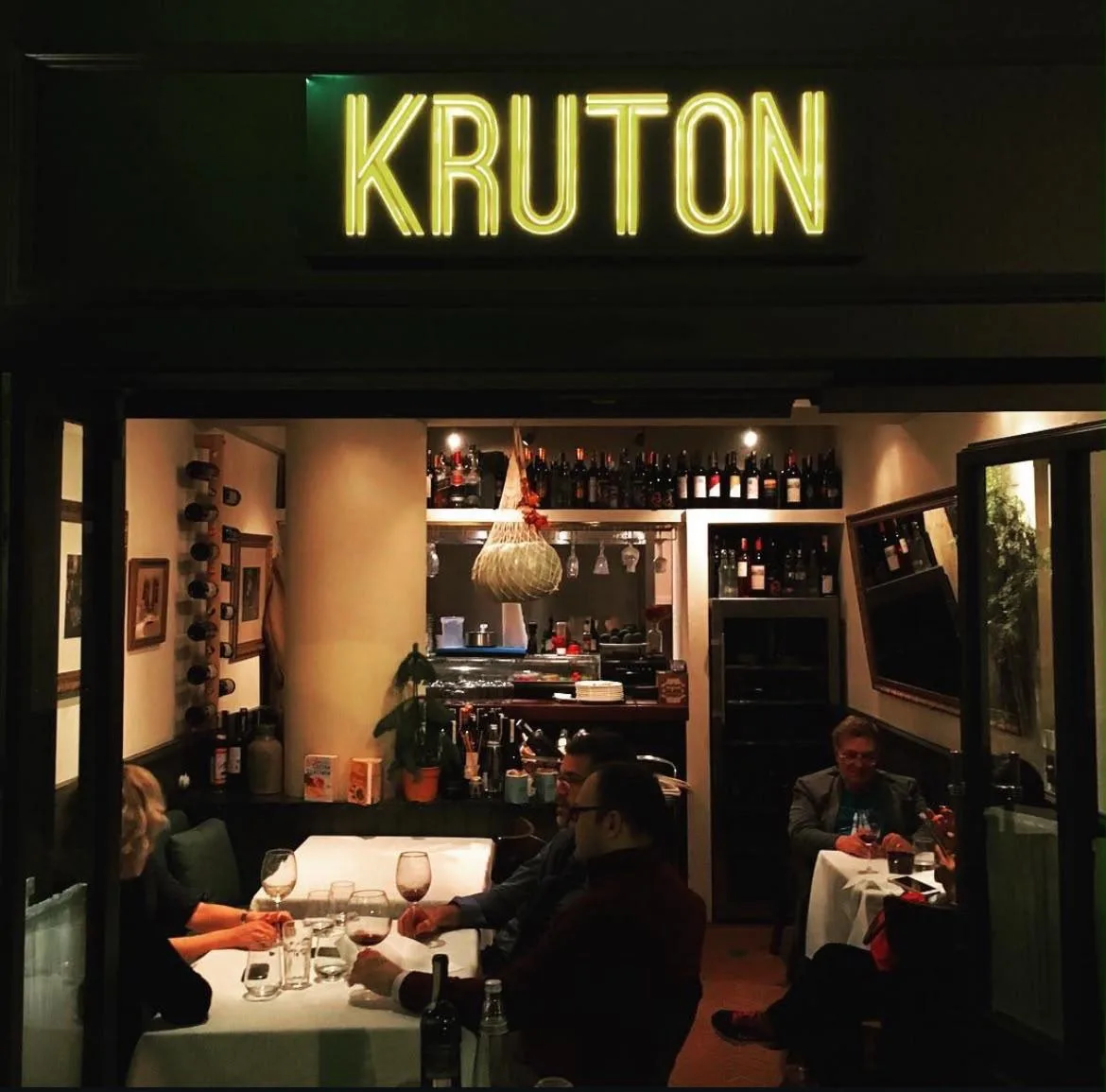 The Kruton sign in yellow neon at night; inside, wine racks, cheeses hanging in netting from the ceiling, and guests at white-clothed tables.