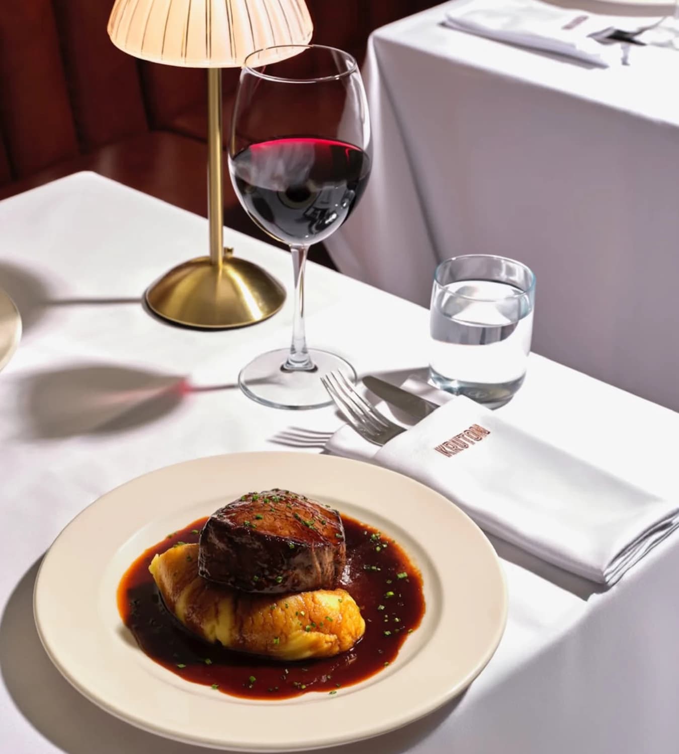 A brass lamp with a pleated shade, a glass of red wine and a KRUTON-embroidered napkin on a white-clothed table; in front, a beef fillet in sauce over puree on a cream plate.