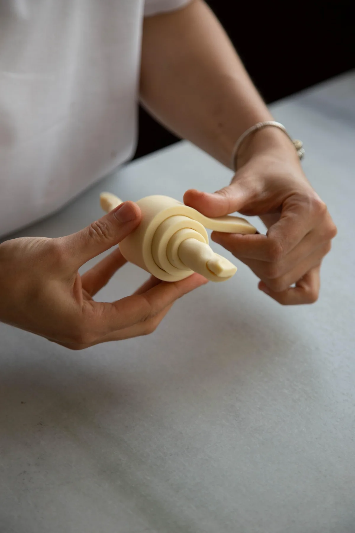 A baker's two hands rolling raw croissant dough end to end on a light-colored counter.