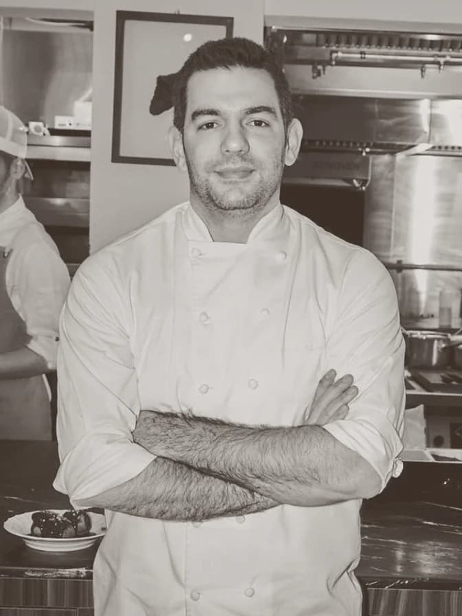 A young chef in a white jacket standing with arms crossed, a stainless steel kitchen in the background.