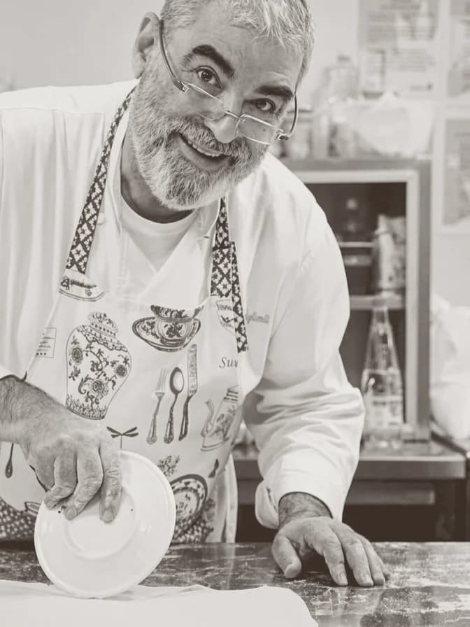 A cook with glasses and a grey beard, a patterned apron over a white jacket, smiling and holding a plate on a marble counter.