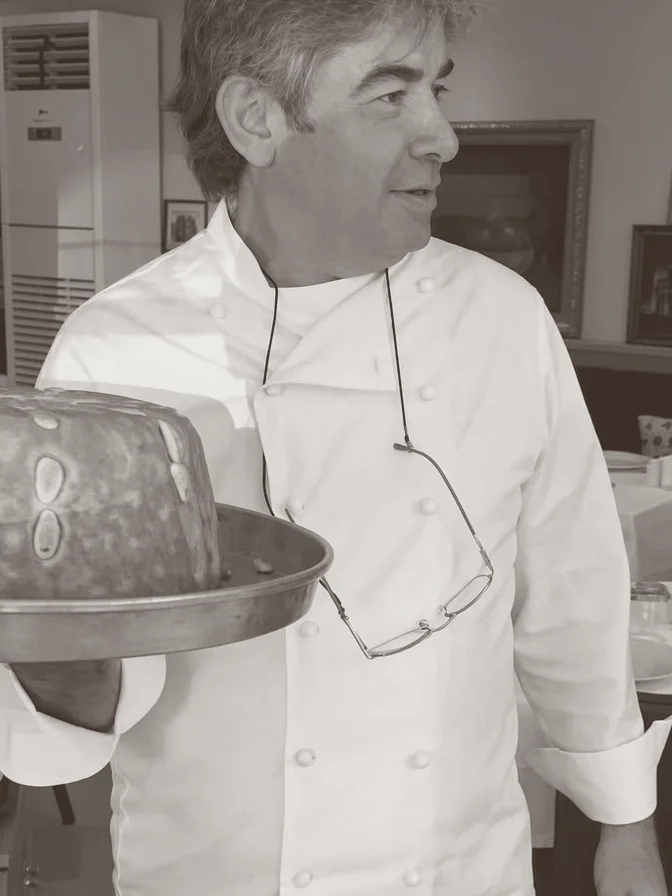 A chef in a white jacket holding a large dessert tray, head turned to the side, glasses hanging on a cord at the neck.