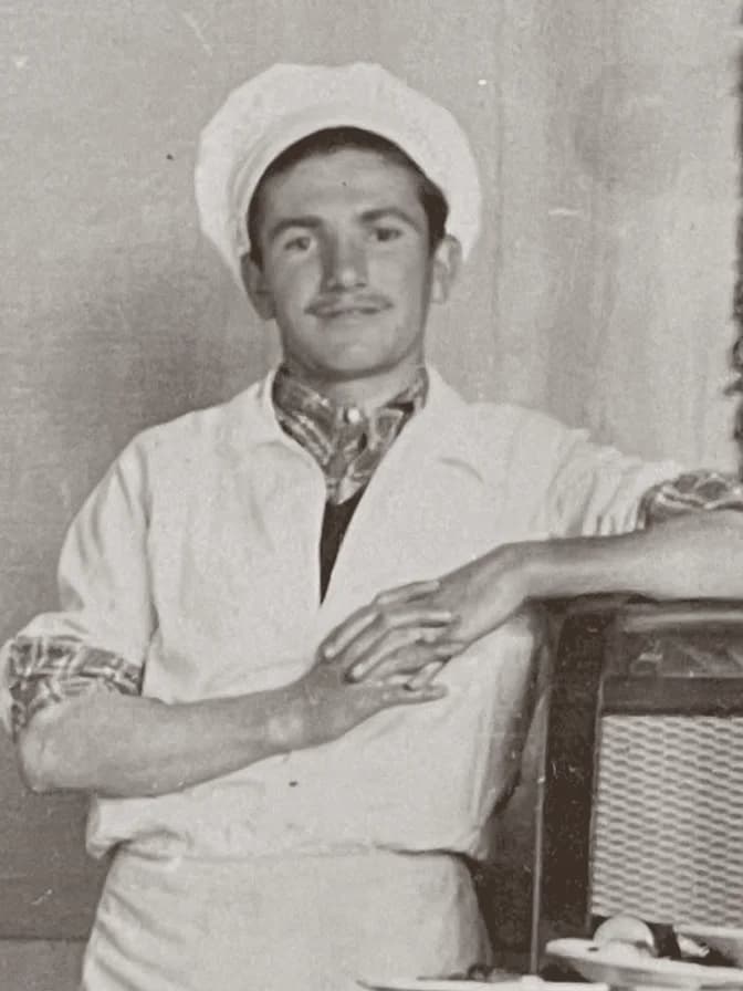 A young cook in an old print: white hat and jacket, a patterned scarf at the neck, leaning an arm on the back of a chair.