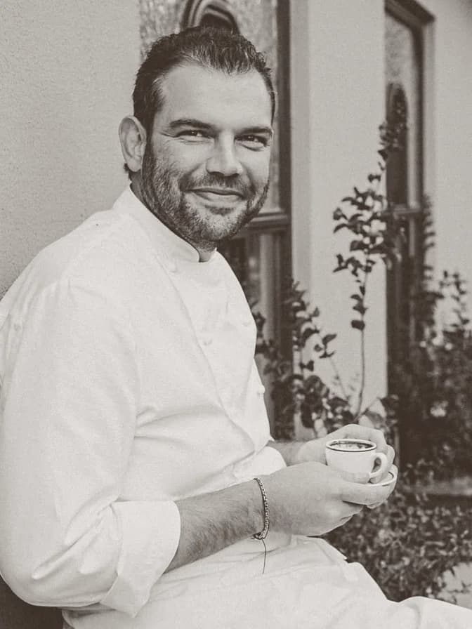 A bearded chef in a white jacket sitting outdoors, holding a coffee cup in both hands and smiling.