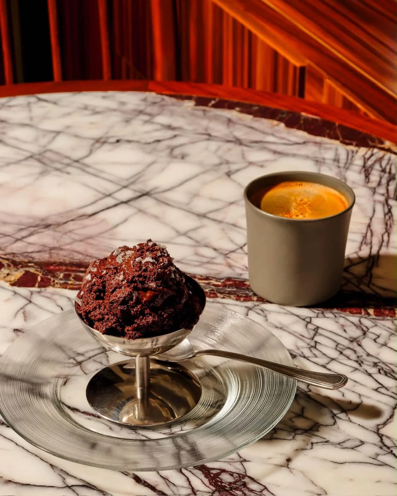 Sea-salted chocolate cookies in a silver bowl beside an espresso on a veined marble table, with wood paneling and a red curtain behind.