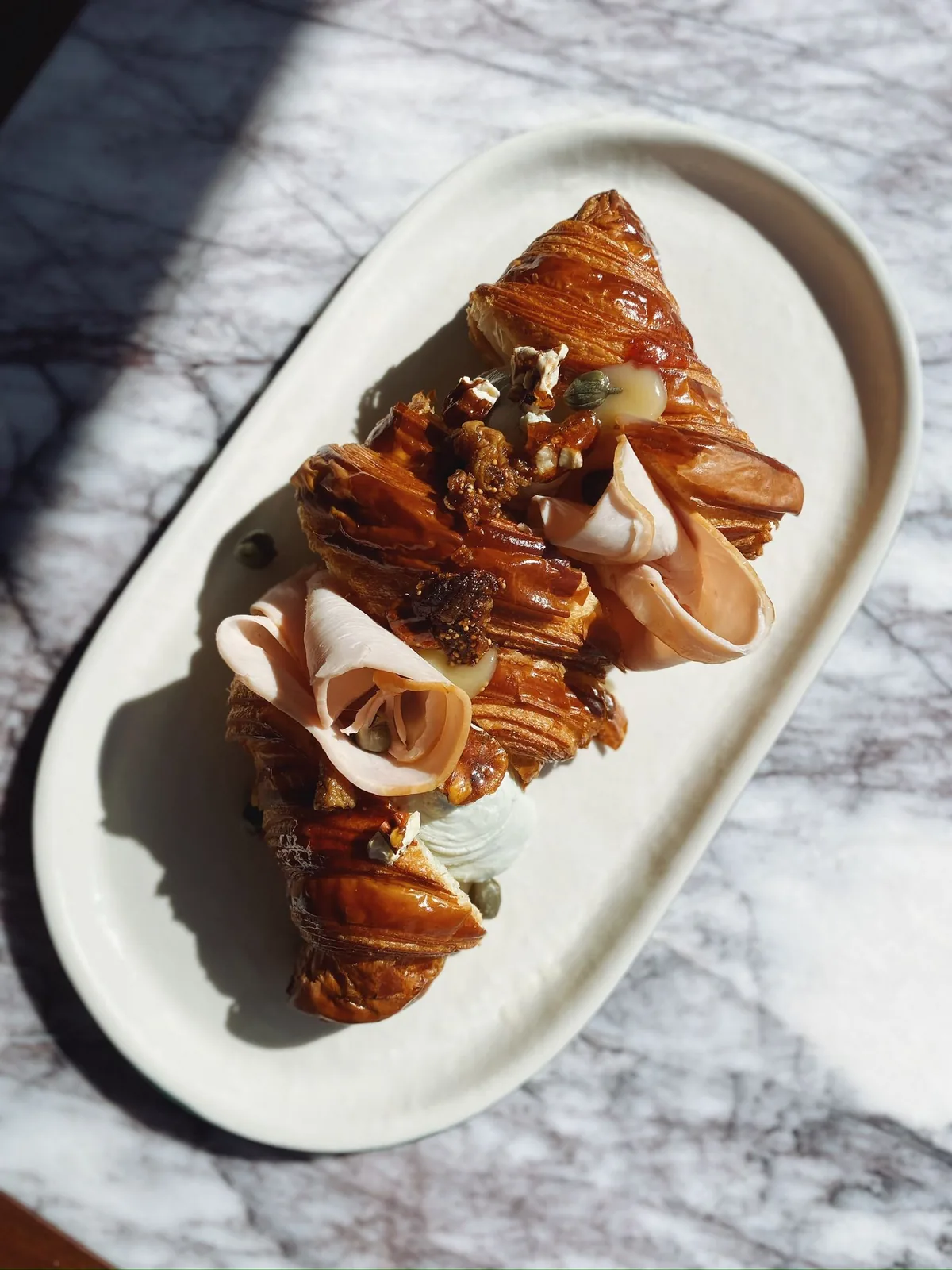 A croissant split open on an oval white plate on a marble table, topped with folded mortadella slices, figs, capers and whipped cheese.