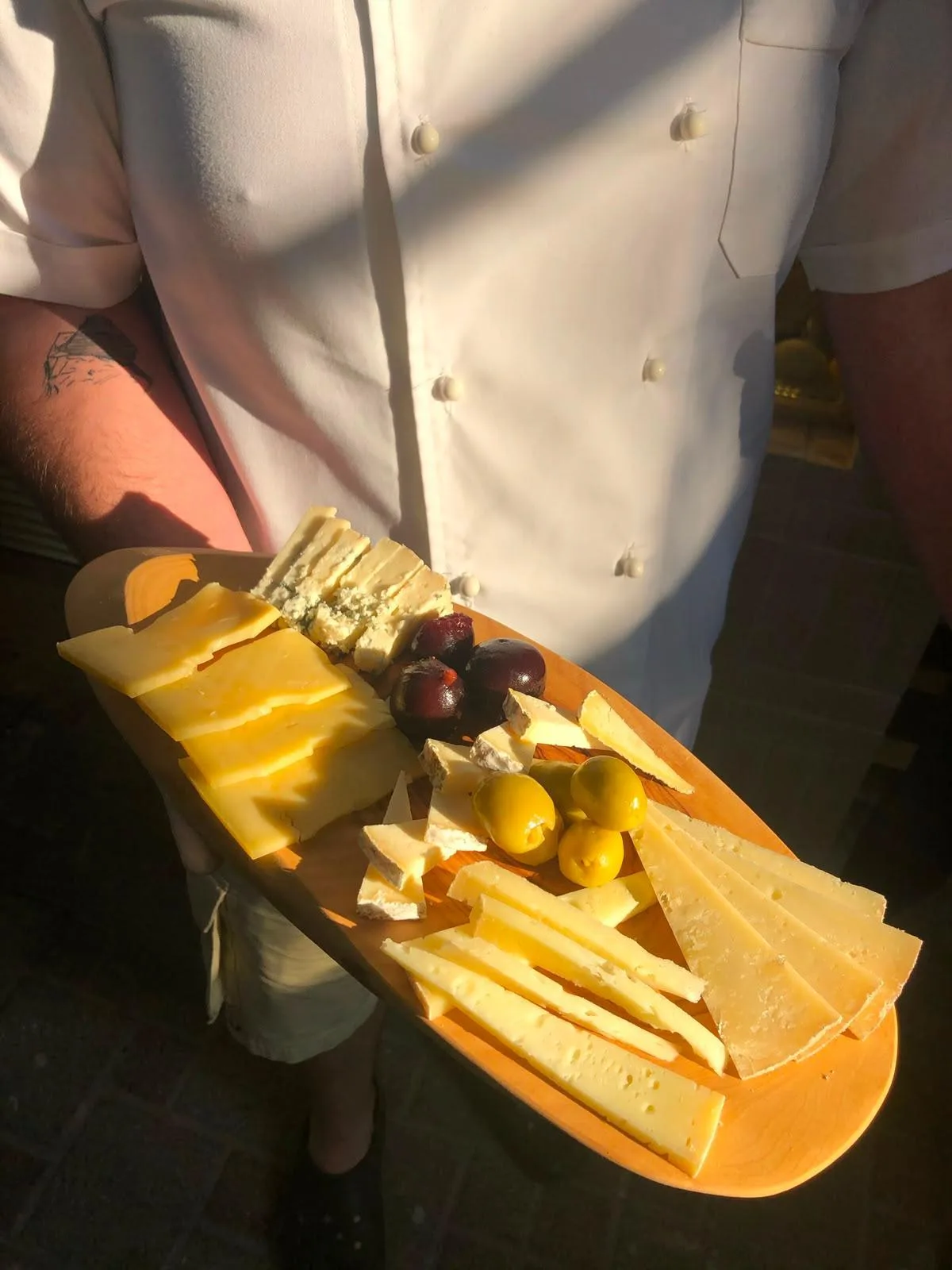 A cook in a white jacket carrying cheeses, olives and dark cherries on an oval wooden board.