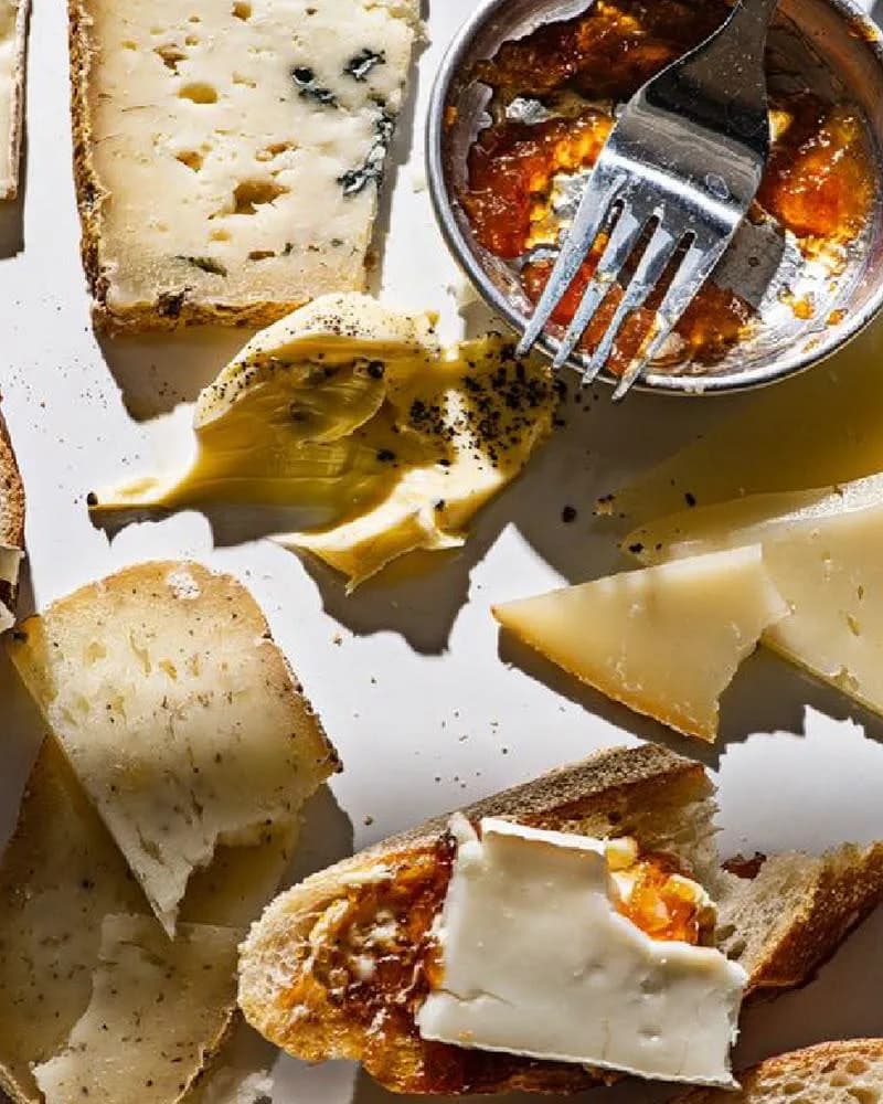 Cheese on toasted bread on a white surface, beside a metal dish of jam with a fork and a slice of brie.