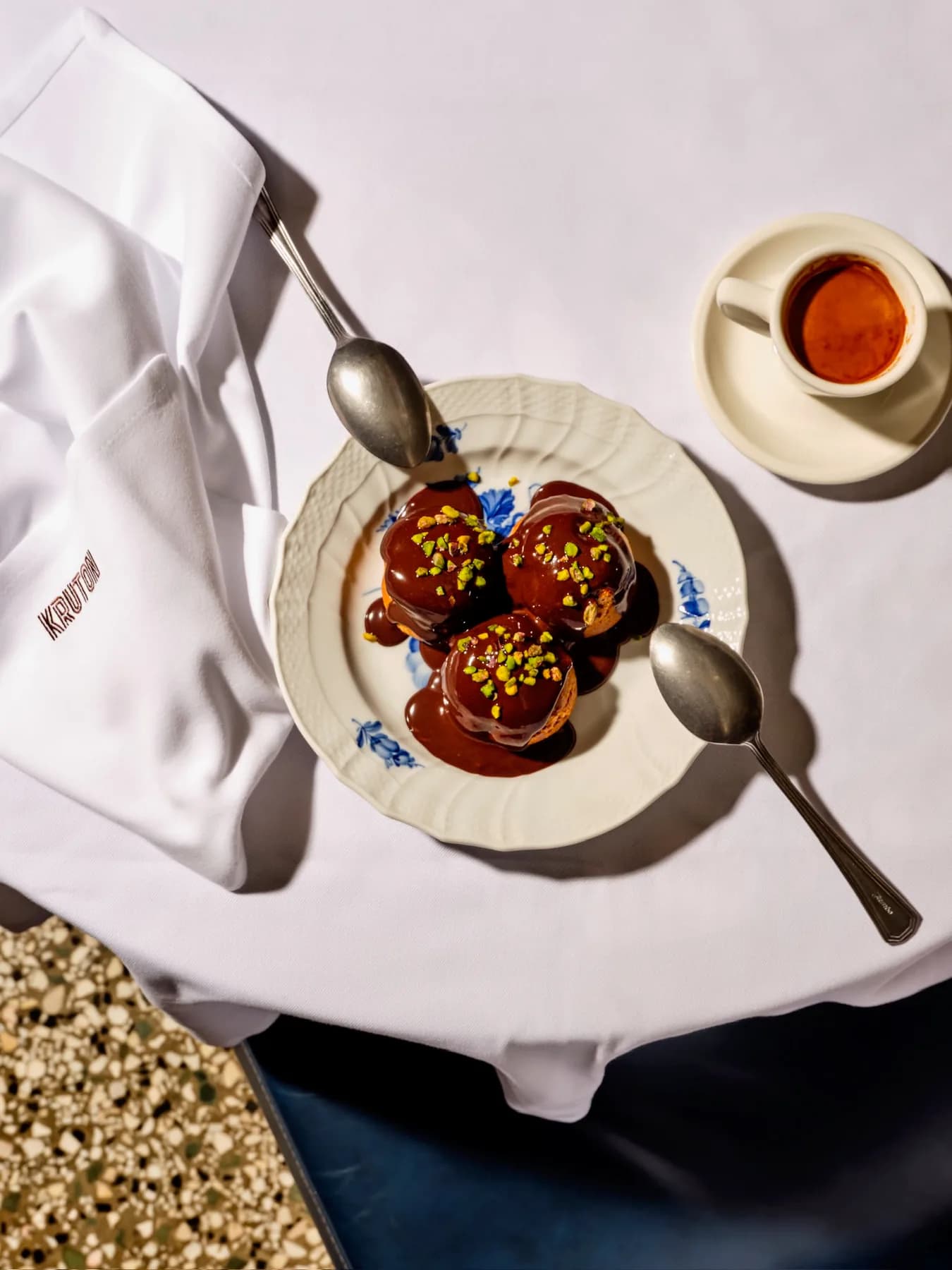 Chocolate-sauced profiteroles beside an espresso, set on a white napkin embroidered with KRUTON.