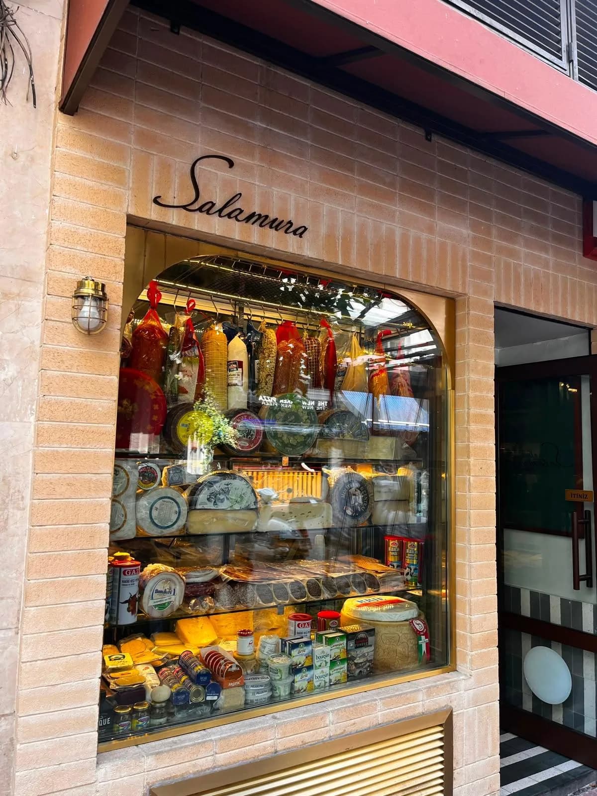 Salamura's storefront: a handwritten sign on a beige brick wall, a lit display case with charcuterie, wheels of cheese and jars.
