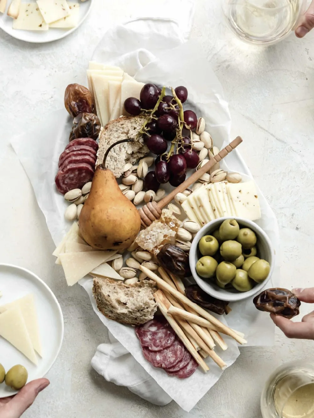 A charcuterie board on a white surface; two hands reaching for the plate, with two glasses of white wine beside it.