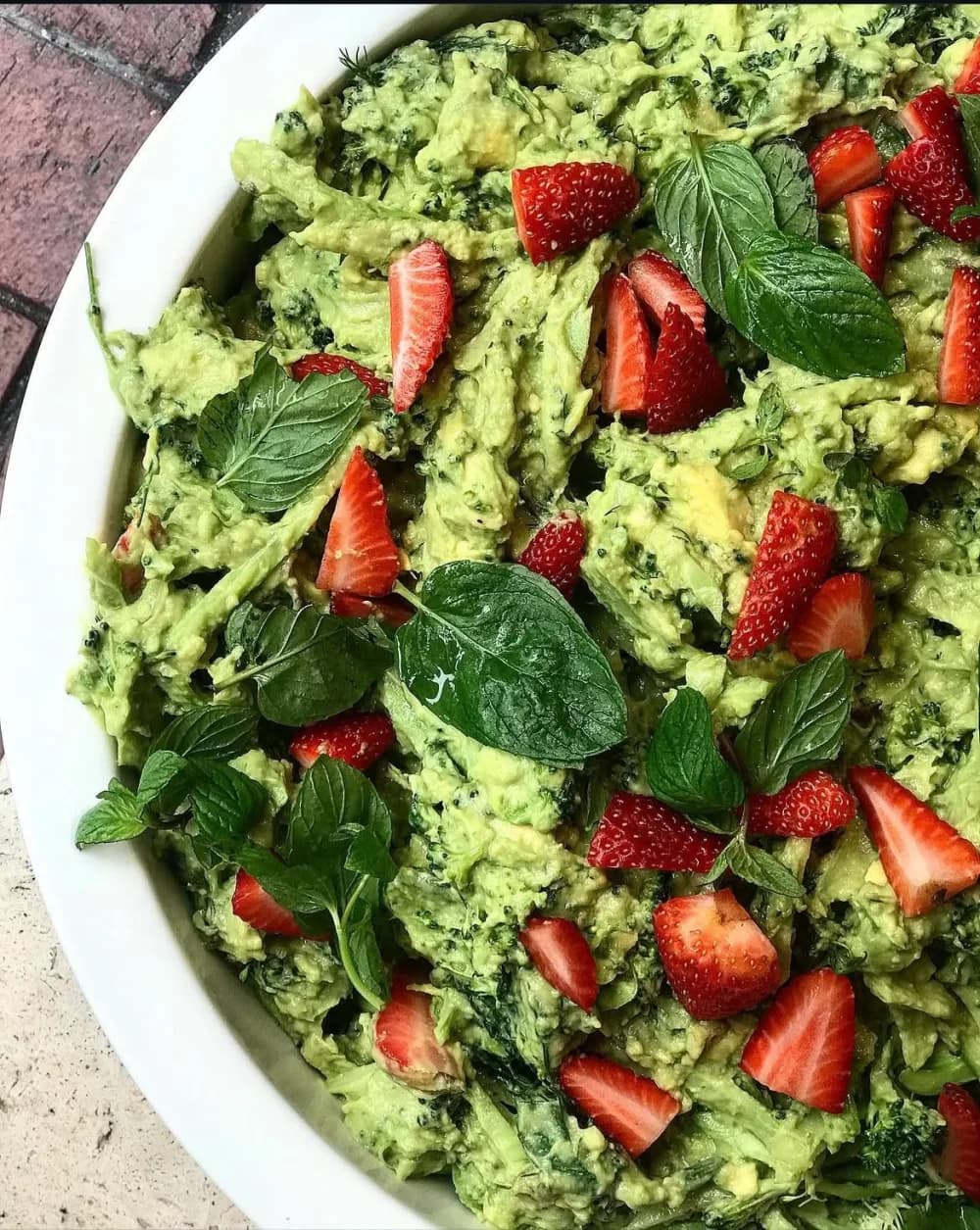 Broccoli and avocado salad in a white bowl, topped with sliced strawberries, mint, basil and dill.